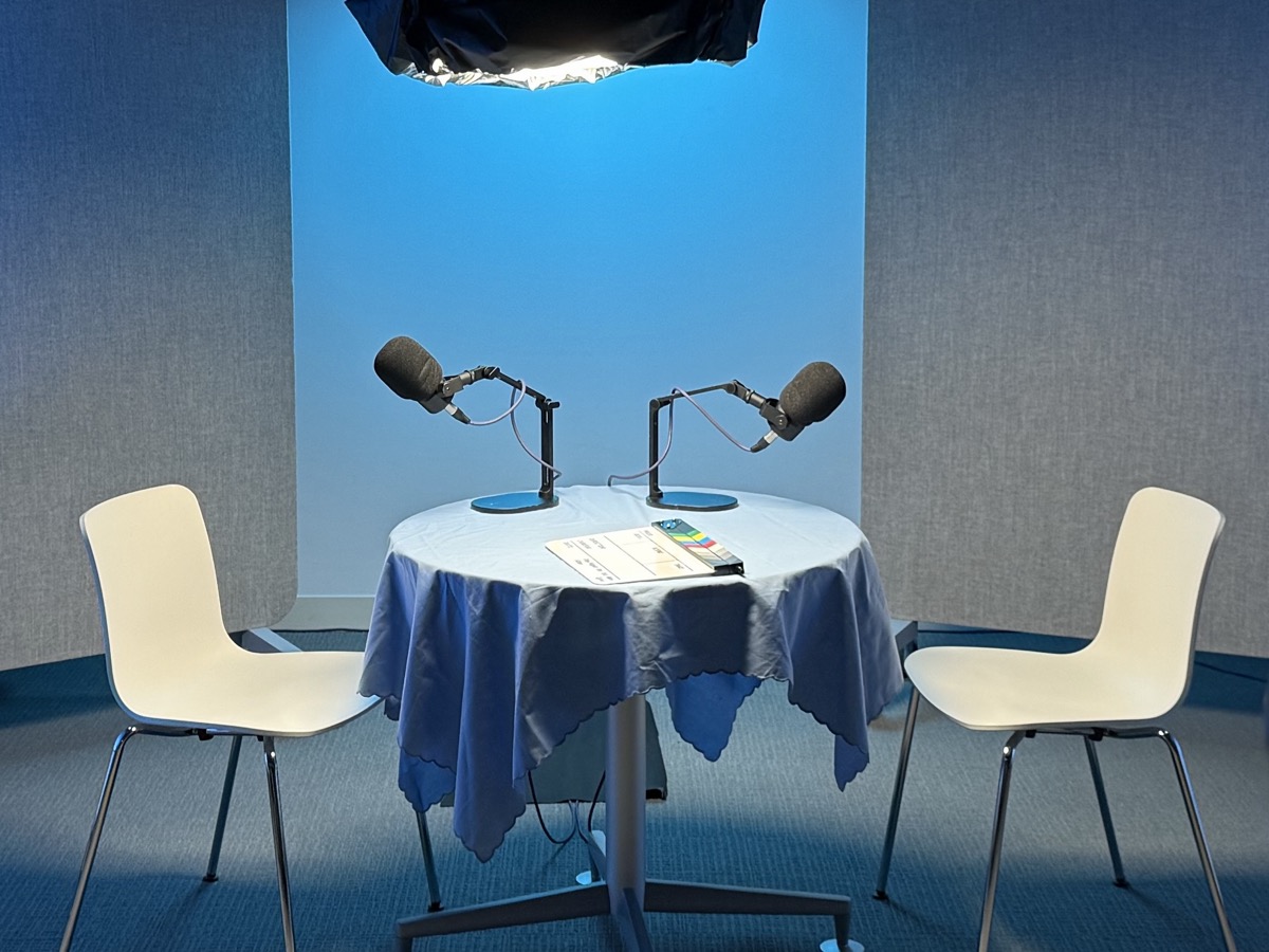A podcast recording table set with two microphones, a clapperboard and two empty chairs, in the Cambridge TV studio